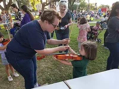 Woman helping a girl play a violin outdoors.